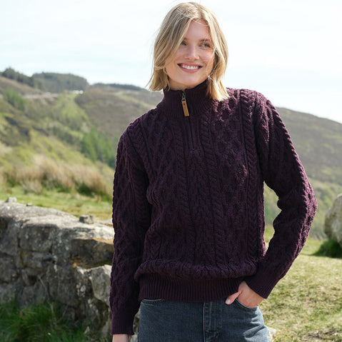 Woman wearing a purple knit sweater standing in a natural landscape with green hills and a stone wall.