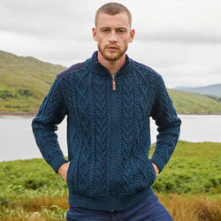 Man wearing a blackwatch blue Men's Fisherman Full-Zip Aran Cardigan standing in a scenic outdoor setting with mountains and water.