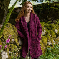 Red headed woman standing by an old stone wall wearing an Irish waterfall Aran cardigan in purple
