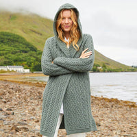 Woman wearing an Irish Aran waterfall cardigan standing by a lake with a pebble shoreline and misty mountains in the background.