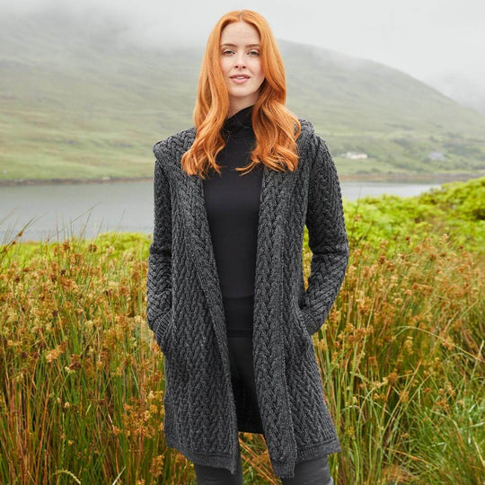 Woman wearing an Irish Aran waterfall cardigan in charcoal gray standing by a lake with lush greenery and misty mountains in the background.