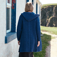 Back view of the Women's Signature Hooded Aran Waterfall Cardigan in Royal Blue, showcasing intricate traditional Irish cable knit stitching and a textured hood, styled beside a traditional Irish cottage along the rugged coastline.