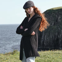 Side view of the Women's Signature Hooded Aran Waterfall Cardigan in Black, featuring traditional Irish cable knit stitching and a relaxed open-front silhouette with hood, styled along the dramatic Irish coastline with rugged sea cliffs in the background.