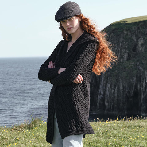 Side view of the Women's Signature Hooded Aran Waterfall Cardigan in Black, featuring traditional Irish cable knit stitching and a relaxed open-front silhouette with hood, styled along the dramatic Irish coastline with rugged sea cliffs in the background.