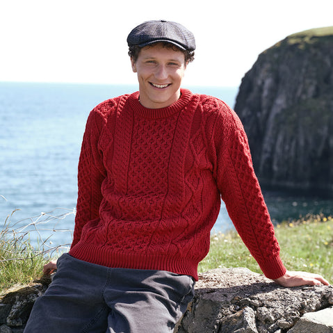 Men's Fisherman Lightweight Crew Neck Heritage Sweater in Cranberry Red, featuring traditional Irish cable knit craftsmanship and heritage-inspired stitching, styled outdoors along the dramatic Irish coastline with rugged sea cliffs in the background.