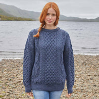 Woman wearing a blue Womens Fisherman Lightweight Crew Neck Heritage Sweater standing on a pebbly beach with water and mountains in the background