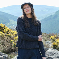 Woman wearing a charcoal Womens Fisherman Lightweight Crew Neck Heritage Sweater and cap standing in a mountainous landscape with yellow flowers.