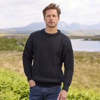 Man wearing a black Men's Fisherman Lightweight Crew Neck Heritage Sweater standing in a scenic outdoor setting with mountains and water.