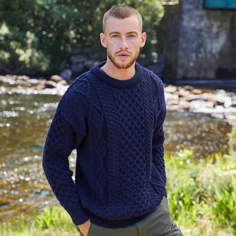 Man wearing a navy Men's Fisherman Lightweight Crew Neck Heritage Sweater standing by a stream with greenery in the background