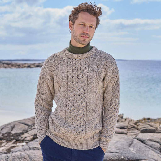 Man wearing a beige Men's Fisherman Lightweight Crew Neck Heritage Sweater standing on a rocky coastal landscape.