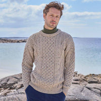 Man wearing a beige Men's Fisherman Lightweight Crew Neck Heritage Sweater standing on a rocky coastal landscape.