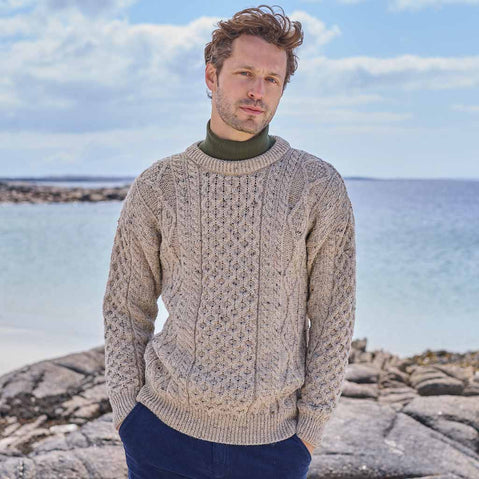 Man wearing a beige Men's Fisherman Lightweight Crew Neck Heritage Sweater standing on a rocky coastal landscape.
