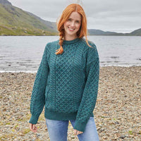 Woman wearing a green Womens Fisherman Lightweight Crew Neck Heritage Sweater standing on a pebbly beach with mountains in the background