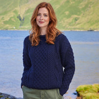 Image featuring the Women's Fisherman Lightweight Crew Neck Heritage Sweater in navy, crafted with authentic Irish fisherman cable knit patterns and a timeless crew neck silhouette, showcasing traditional Irish craftsmanship against a scenic lakeside landscape inspired by Ireland's rugged countryside.