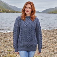 Image featuring the Women's Fisherman Lightweight Crew Neck Heritage Sweater in blue, crafted with authentic Irish fisherman cable knit patterns and a timeless crew neck silhouette, showcasing traditional Irish craftsmanship against a scenic lakeside landscape inspired by Ireland's rugged countryside.