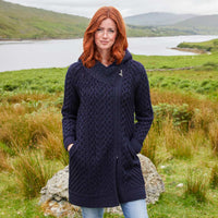 Image featuring the Women's Heritage Aran Hooded Zipper Coat in navy, crafted with authentic Irish Aran cable knit patterns, a practical hood, full-zip closure, and spacious front pockets, showcasing timeless Irish craftsmanship against a scenic lakeside landscape inspired by Ireland's rugged countryside.