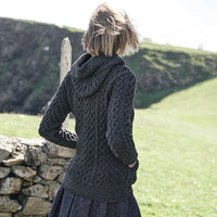 Back view of the Women's Signature Aran Celtic Zipper Hooded Cardigan in Charcoal, showcasing intricate traditional Irish cable knit patterns and a textured hood, styled beside a rustic stone wall with the rolling Irish countryside in the background.