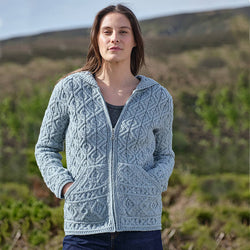 Woman wearing a mermaid-colored Women's Signature Aran Zipper Hoodie from The Irish Store, featuring intricate cable knit patterns and a front zipper closure, standing outdoors with a blurred background of greenery and hills.