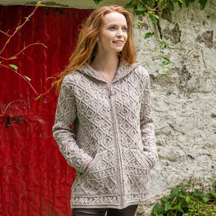 Woman wearing an oatmeal Women's Signature Aran Zipper Hoodie standing against a red wall and stone wall.