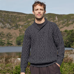 Man wearing a dark gray sweater standing in a natural landscape with mountains and water.