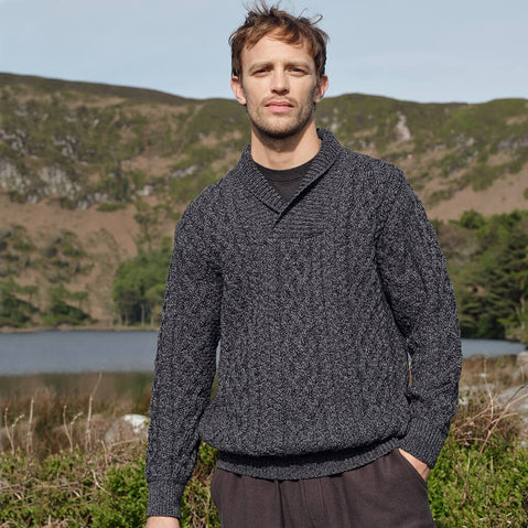 Man wearing a dark gray sweater standing in a natural landscape with mountains and water.