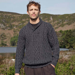 Man wearing a charcoal Men's Shawl Collar Heritage Aran Sweater standing in a natural landscape with mountains and water.