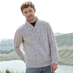 Man wearing a cable knit sweater standing in front of a scenic landscape with mountains and water.