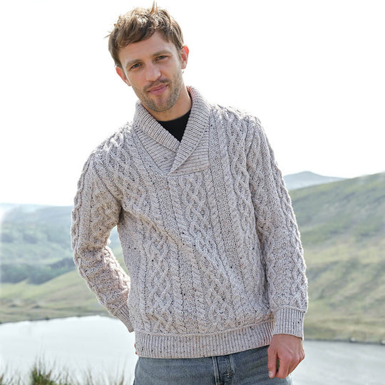 Man wearing a cable knit sweater standing in front of a scenic landscape with mountains and water.