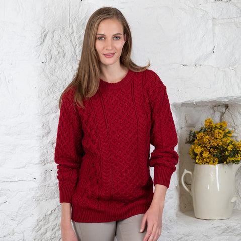A woman standing by the water wearing a red Irish wool crew neck sweater with a green scarf and a smile on her face.