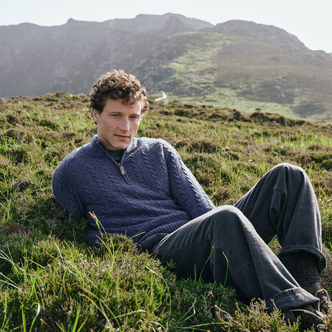 Image featuring the Men's Traditional Half Zip Cable Knit Sweater in indigo, crafted with authentic Irish cable knit patterns and a practical half-zip neckline, showcased against Ireland-inspired mountain scenery.