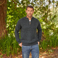 A man standing in a lush green forest wearing a dark gray colored half-zip Irish sweater with a plain stitch design.