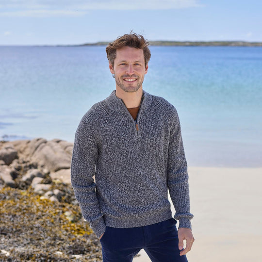 A man standing on a beach wearing a gray half-zip Irish sweater with a plain stitch design.