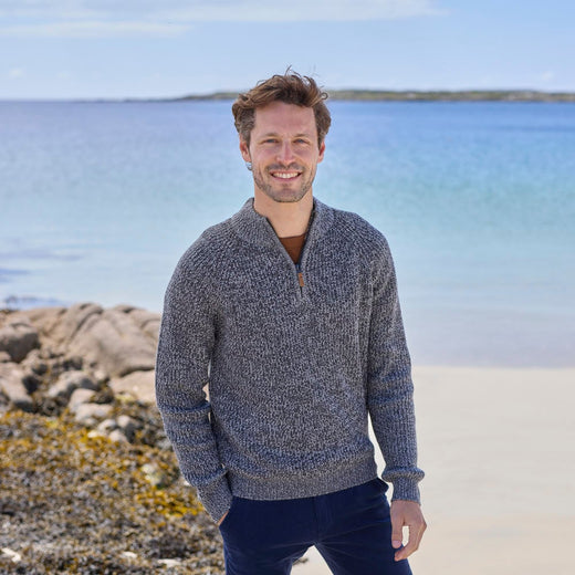 A man standing on a beach wearing a gray half-zip Irish sweater with a plain stitch design.