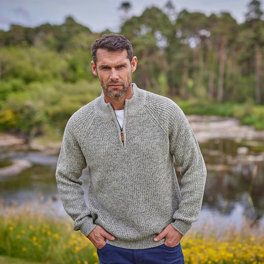 A man standing by a lake wearing an oatmeal colored half-zip Irish sweater with a plain stitch design.