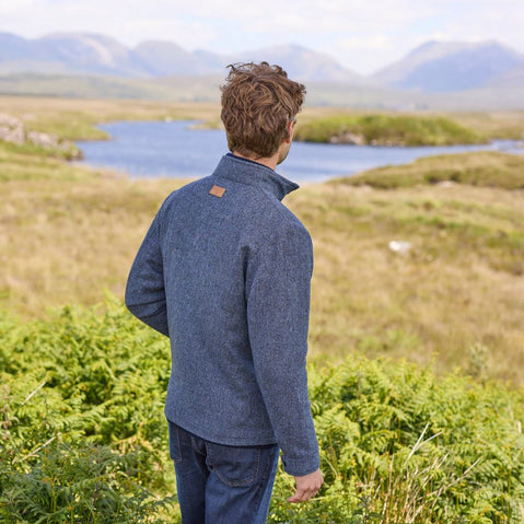 A man standing outdoors wearing a blue Donegal herringbone zipper jacket with long sleeves, paired with jeans.