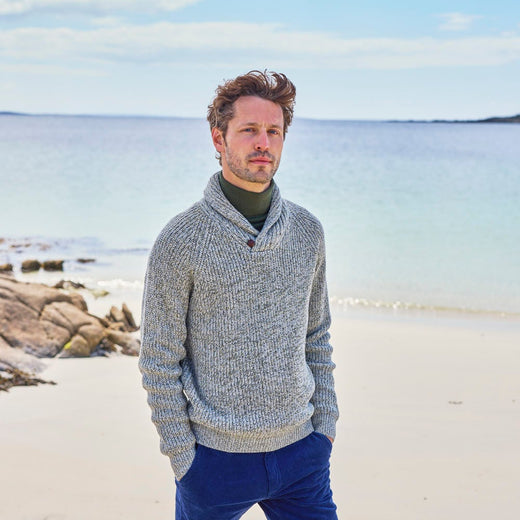 A man standing on a beach wearing a gray shawl collar button sweater and blue trousers.