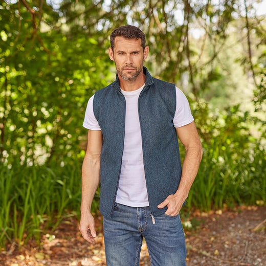 A man wearing a blue tweed Irish windbreaker vest with a white t-shirt and blue jeans, standing in a natural outdoor setting.