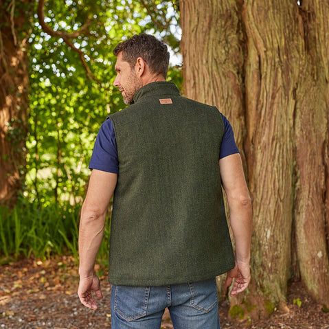 A man wearing a blue tweed Irish windbreaker vest with a white t-shirt and blue jeans, standing in a natural outdoor setting.