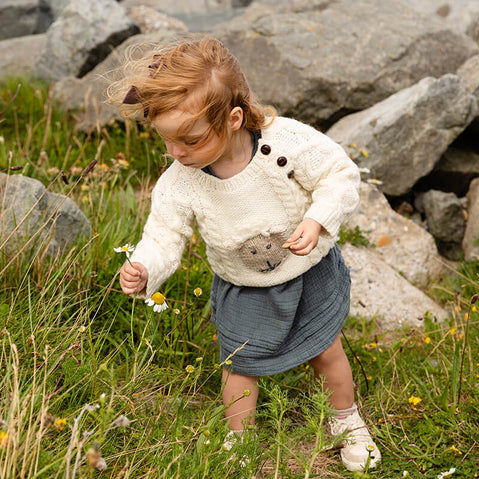 A young child with curly hair is picking flowers in a grassy area surrounded by rocks. She is wearing a cream-colored kids Aran sweater with a bear design and a blue skirt. The scene captures a playful moment outdoors, emphasizing the child's interaction with nature.