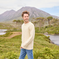 A man standing outdoors wearing a beige hand knit Aran sweater with a mountain and trees in the background.
