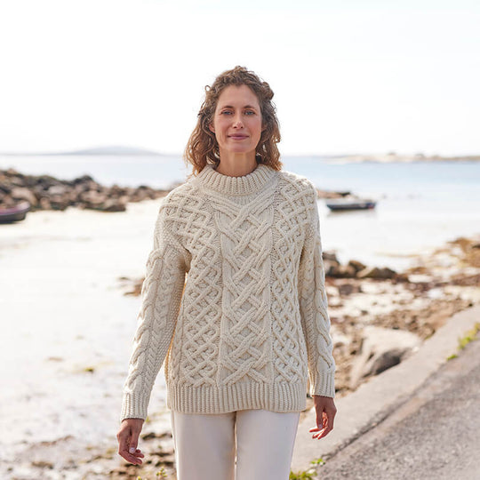 A woman standing by the sea, wearing a beige Aran sweater with a trellis pattern.