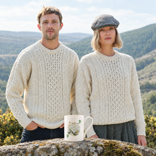 A man and a woman standing outdoors in front of a mountain landscape, both wearing chunky cream cable-knit crewneck sweaters. A white ceramic mug with a botanical illustration and text sits on a mossy stone in the foreground.