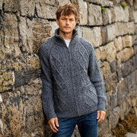A man stands against a stone wall, wearing a gray knitted aran sweater with a zipper and ribbed collar. He has tousled hair and is looking directly at the camera, displaying a relaxed pose. The background features rustic stonework, suggesting an outdoor setting.