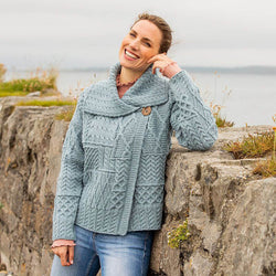 A woman stands against a stone wall by the water, smiling and wearing a light blue knitted aran sweater with a shawl collar. She has long hair and is casually posing, with lush greenery in the background. The scene conveys a relaxed outdoor atmosphere.