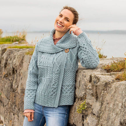 A woman stands against a stone wall by the water, smiling and wearing a light blue knitted aran sweater with a shawl collar. She has long hair and is casually posing, with lush greenery in the background. The scene conveys a relaxed outdoor atmosphere.