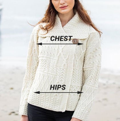 A woman poses on the beach wearing a textured white aran sweater with a shawl collar. Labels indicating 
