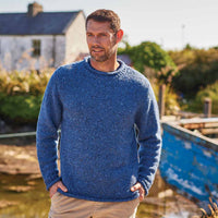 Man wearing a blue Men's Traditional Fisherman Irish Wool Sweater standing outdoors with a house and boat in the background