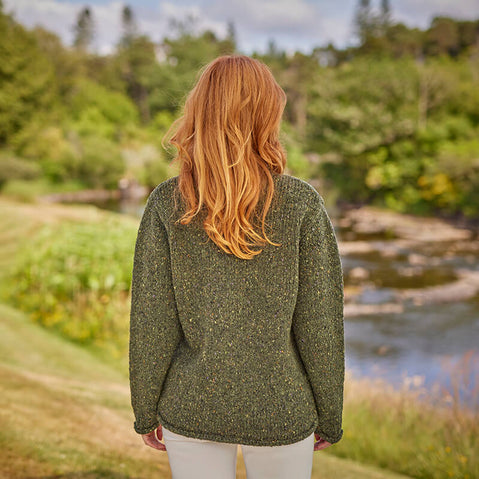 A woman standing outdoors wearing a green flecked Irish wool sweater with a round neckline, long sleeves, and casual fit.