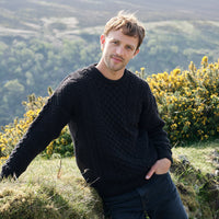 Man wearing a black sweater standing in a natural setting with greenery and yellow flowers.