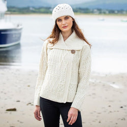 A woman stands on a beach, wearing a knitted cream-colored aran sweater with textured patterns and a button detail. She also wears a matching knit hat. The background features a calm sea with a boat nearby and mountains in the distance.
